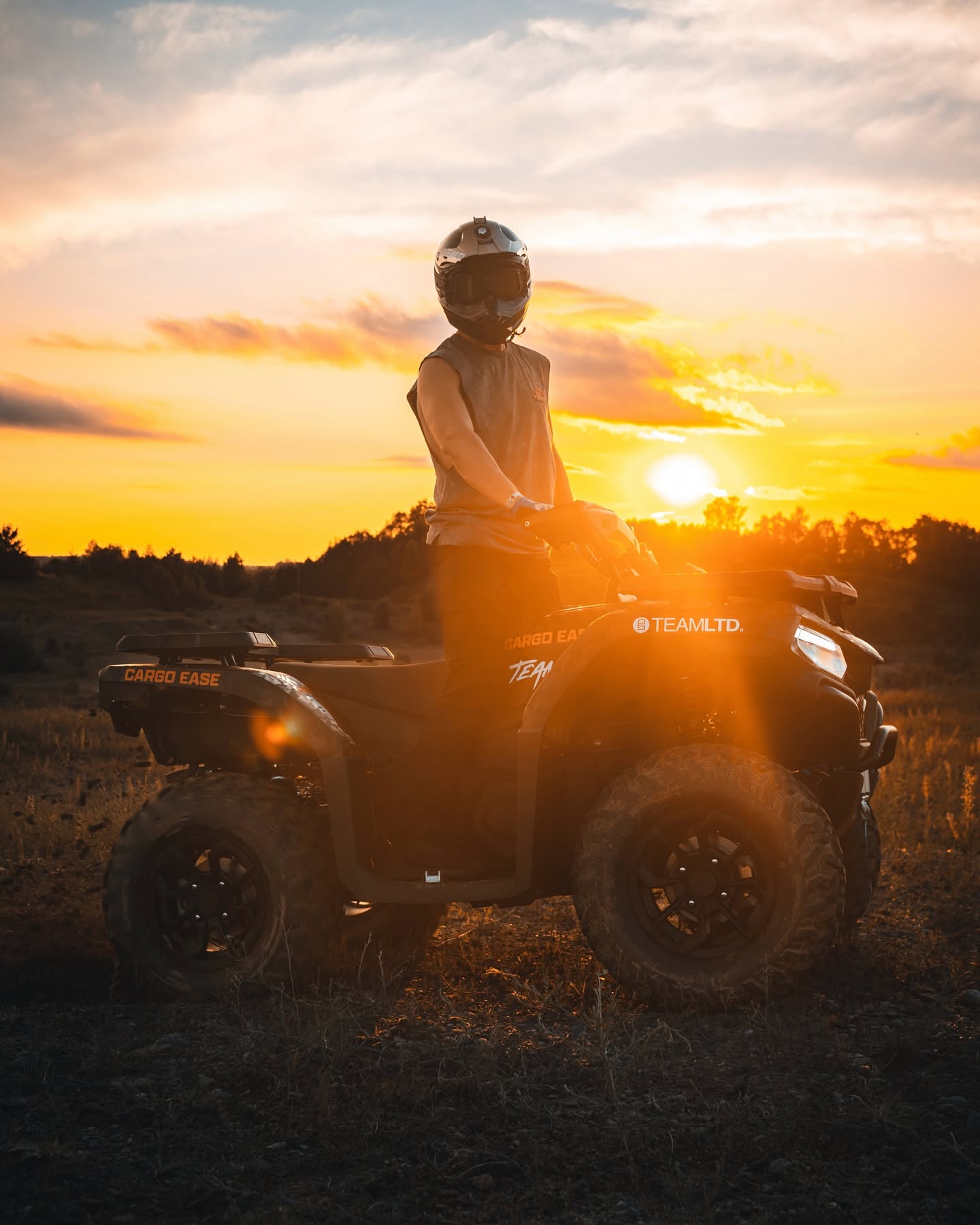 A person wearing a helmet sits on a black all-terrain vehicle (ATV) in an open field at sunset, with warm golden light illuminating the scene and the ATV.